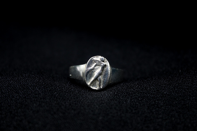 Pu&ograve; includere: Un anello d'argento con un designo in rilievo di una donna con un vestito fluido. L'anello &egrave; su uno sfondo nero.