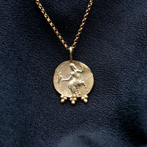 May include: A gold chain necklace with a round pendant featuring a detailed image of a woman holding a bird. The pendant has a textured surface and is accented with small gold beads.