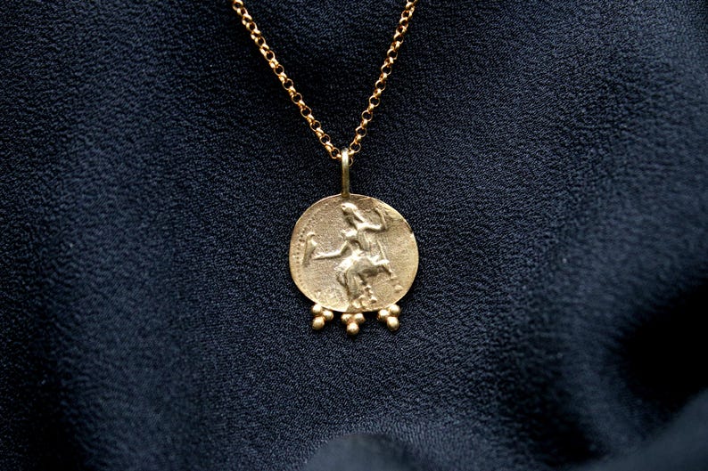 May include: A gold chain necklace with a round pendant featuring a detailed image of a woman holding a bird. The pendant has a textured surface and is accented with small gold beads.