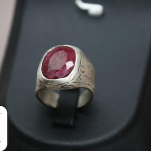 May include: A silver ring with a large, oval-shaped, red ruby gemstone. The ring band is engraved with intricate designs.