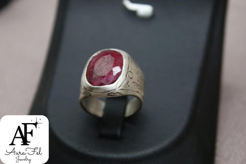 May include: A silver ring with a large, oval-shaped, red ruby gemstone. The ring band is engraved with intricate designs.