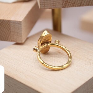 May include: A gold ring with a hammered texture and a large, irregular-shaped stone. The ring is on a wooden surface.