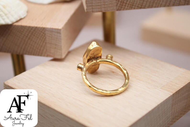 May include: A gold ring with a hammered texture and a large, irregular-shaped stone. The ring is on a wooden surface.