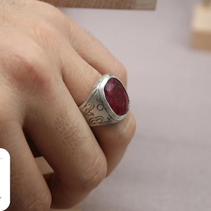 May include: A silver ring with a large, oval-shaped, red gemstone set in the center. The ring band is engraved with intricate designs.