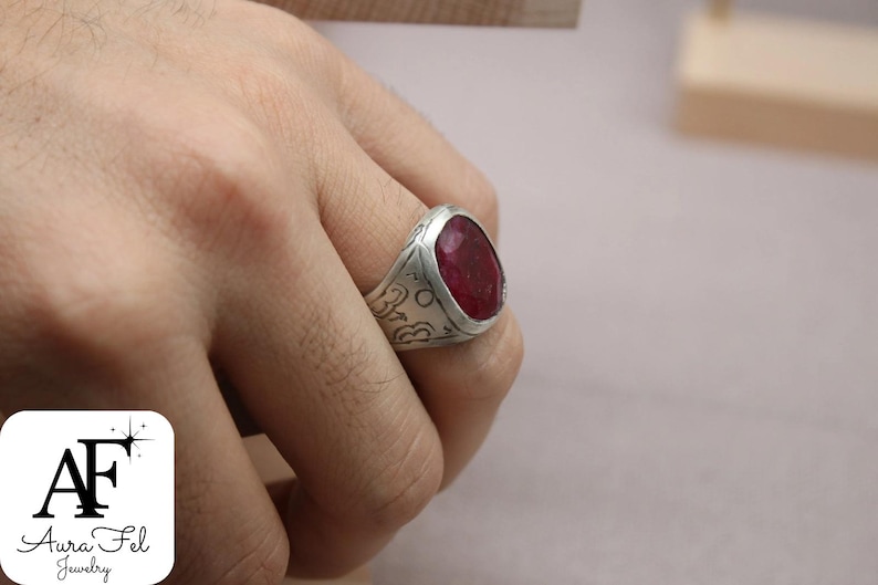 May include: A silver ring with a large, oval-shaped, red gemstone set in the center. The ring band is engraved with intricate designs.