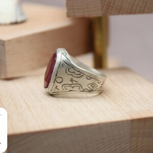 May include: A silver ring with a red gemstone and intricate engraved details. The ring is displayed on a wooden surface.