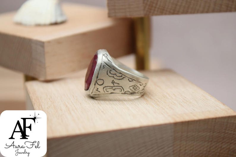 May include: A silver ring with a red gemstone and intricate engraved details. The ring is displayed on a wooden surface.