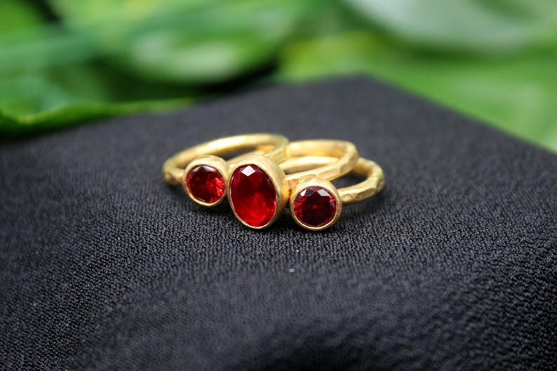 May include: Three gold rings with red oval-shaped gemstones. The rings are stacked on top of each other.