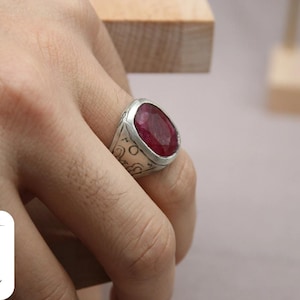 May include: A silver ring with a large, oval-shaped, deep red gemstone set in the center. The ring has intricate designs engraved on the band.