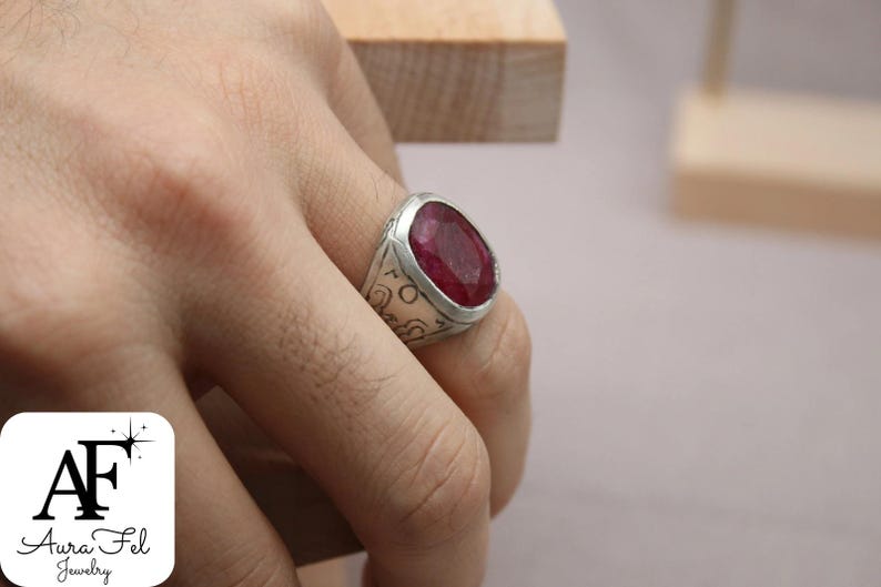 May include: A silver ring with a large, oval-shaped, deep red gemstone set in the center. The ring has intricate designs engraved on the band.