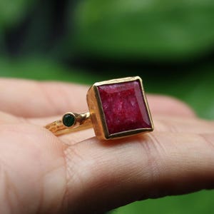 May include: A gold ring featuring a large, square, red ruby gemstone and a smaller, round, green gemstone.