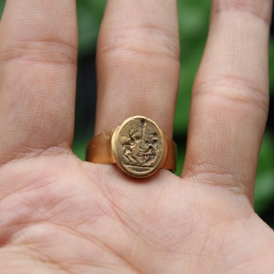 May include: A gold ring with a raised oval design featuring a detailed image of a person riding a horse. The ring is worn on a person's finger.