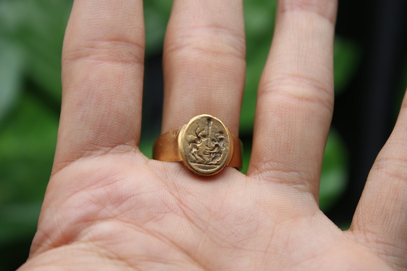 May include: A gold ring with a raised oval design featuring a detailed image of a person riding a horse. The ring is worn on a person's finger.