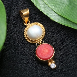 Op de afbeelding: Gouden hanger met een witte parel en een rode koraalsteen, verbonden door een gouden ketting. De hanger heeft een klein diamantaccent aan de onderkant. De sieraden zijn geplaatst tegen een zwarte achtergrond.