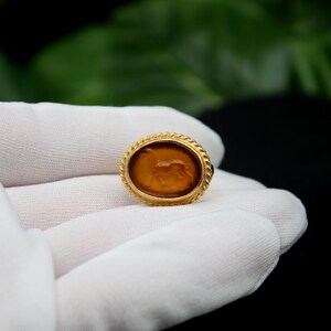 May include: A gold ring with a brown oval cameo featuring a carved image of a lion. The ring is on a white surface.