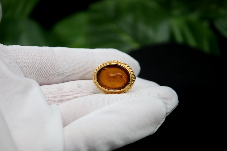 May include: A gold ring with a brown oval cameo featuring a carved image of a lion. The ring is on a white surface.