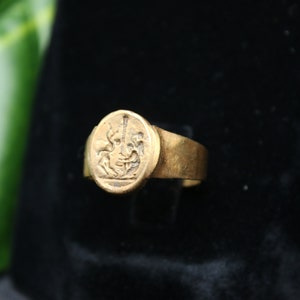 May include: A gold ring with a raised oval design featuring a detailed image of a mythical creature. The ring is set against a black background.