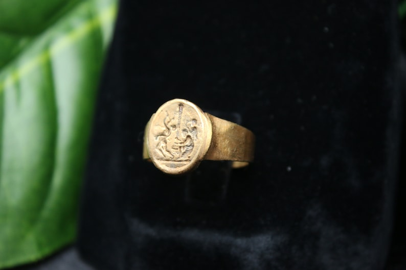 May include: A gold ring with a raised oval design featuring a detailed image of a mythical creature. The ring is set against a black background.