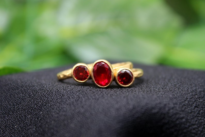 May include: A gold ring with three red gemstones. The ring is on a black fabric background.