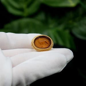 May include: A gold-colored ring with a brown oval cameo featuring a carved image of a lion. The ring is on a white surface.