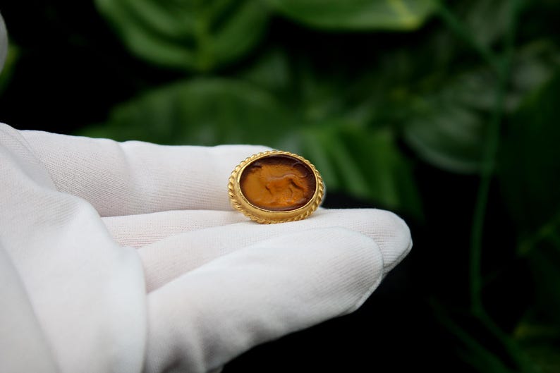May include: A gold-colored ring with a brown oval cameo featuring a carved image of a lion. The ring is on a white surface.