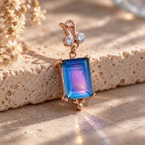 May include: A rose gold pendant featuring a rectangular iridescent blue and purple gemstone. The setting includes two small clear round stones at the top bail and a small metal bead at the bottom. The pendant has a polished metallic finish.