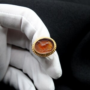 May include: A gold ring with a carved carnelian stone featuring a detailed depiction of a running animal. The stone is set in a gold bezel with a decorative border.