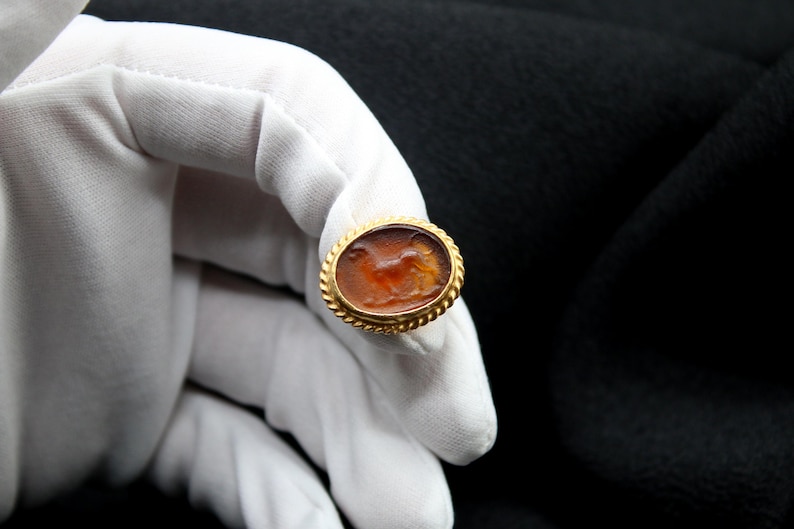 May include: A gold ring with a carved carnelian stone featuring a detailed depiction of a running animal. The stone is set in a gold bezel with a decorative border.