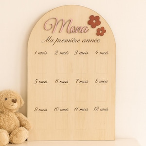 Peut inclure: Tableau de jalons pour bébé en bois avec le nom "Mona" en écriture rose et deux fleurs rouges. Le tableau comporte des espaces pour les photos de 1 à 12 mois, avec le texte "Ma première année" au-dessus des emplacements photo.