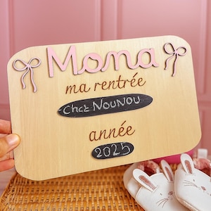 Op de afbeelding: Houten bord met roze tekst en strikaccenten. Het bord leest "Mona, ma rentrée, Chez NouNou, année 2025". Het bord wordt door een hand vastgehouden, met een roze achtergrond.