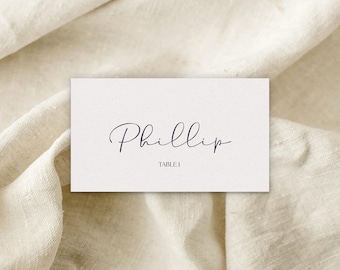 Pode incluir: Um cartão de lugar retangular com o nome "Phillip" em elegante escrita cursiva. Abaixo do nome está o texto "TABLE 1". O cartão está colocado sobre um fundo de tecido neutro e texturizado.
