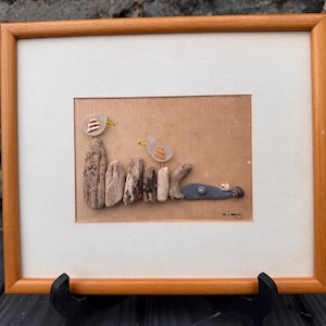 Puede incluir: Obra de arte enmarcada que representa una escena de playa con dos pájaros y madera flotante. Los pájaros están hechos de vidrio transparente con picos amarillos. Las piezas de madera flotante están dispuestas para crear un paisaje. El marco es de madera.