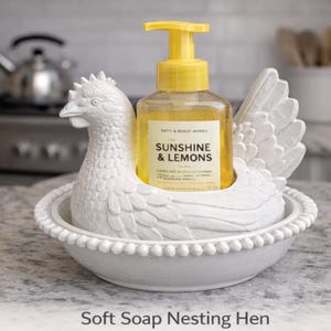 K&ouml;nnte beinhalten: Ein wei&szlig;er Keramik-Seifenspender in Form eines Huhns h&auml;lt eine klare Flasche Fl&uuml;ssigseife mit gelbem Pumpkopf. Auf der Seifenflasche steht "Sunshine & Lemons". Der Spender steht auf einer Arbeitsplatte.