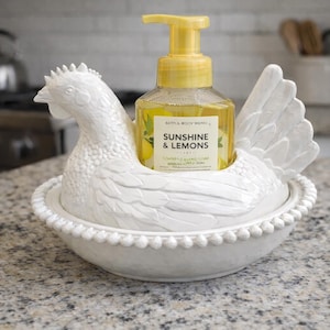 Pode incluir: Um suporte de dispensador de sabonete em forma de galinha de cer&acirc;mica branca com uma garrafa de sabonete l&iacute;quido amarelo. A garrafa de sabonete tem uma bomba amarela e um r&oacute;tulo que diz "Sunshine & Lemons". O suporte est&aacute; em uma bancada salpicada.