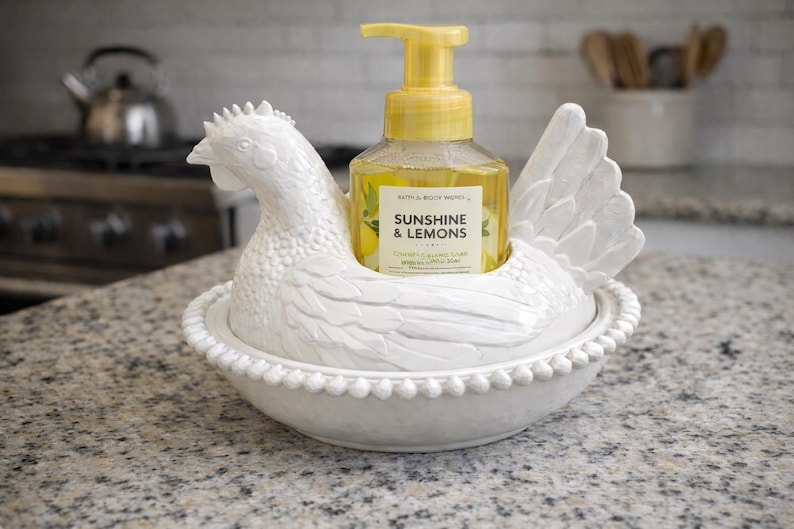 Pode incluir: Um suporte de dispensador de sabonete em forma de galinha de cer&acirc;mica branca com uma garrafa de sabonete l&iacute;quido amarelo. A garrafa de sabonete tem uma bomba amarela e um r&oacute;tulo que diz "Sunshine & Lemons". O suporte est&aacute; em uma bancada salpicada.