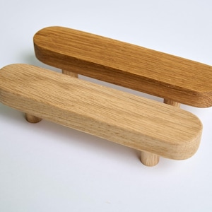 Könnte beinhalten: Zwei Holzknaufgriffe mit abgerundeter ovaler Form und einem zylindrischen Knopf. Die Griffe sind aus hellem braunem Holz und unbehandelt.