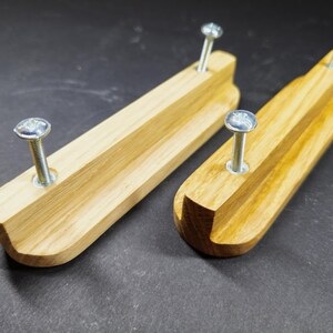 Op de afbeelding: Twee lichtgekleurde houten ladehandgrepen met een gladde, langwerpige rechthoekige vorm en een gebogen rand. Elke handgreep is voorzien van twee zilverkleurige metalen schroeven die door het hout zijn gestoken en naar boven uitsteken.
