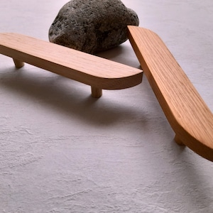 May include: Two wooden drawer pulls with rounded edges. The pulls are made of light brown wood and have a simple, modern design.