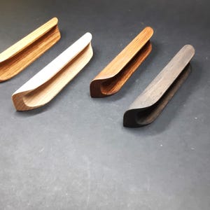 May include: Four wooden cabinet pulls in different wood finishes: natural, light wood, dark wood, and black. Each pull has a curved, rectangular shape with a recessed centre.
