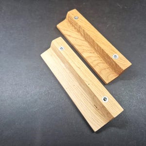 May include: Two rectangular light brown wooden cabinet pulls with a natural wood grain pattern. Each handle features an L-shaped profile and is secured with two silver-coloured metal screws positioned along the top edge of the wooden surface.