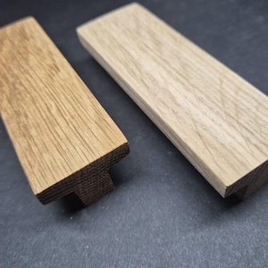 May include: Two rectangular wooden cabinet pulls with a T-shaped profile. One pull features a medium brown wood grain finish, while the other pull displays a lighter, pale beige wood grain finish. Both items have flat tops and angled sides.