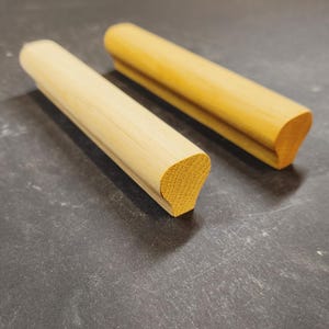 May include: Two elongated wooden handrail components with a rounded top profile and a flat base. One piece features a light, natural wood grain finish, while the other displays a slightly darker, warmer golden-yellow wood tone and texture.