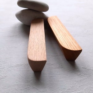 Peut inclure: Deux poignées de tiroir en bois avec une forme arrondie légèrement incurvée. Les poignées sont en bois brun clair avec un motif de grain naturel.