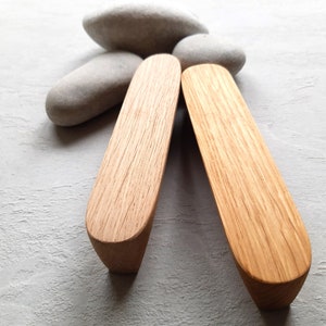 May include: Two wooden cabinet pulls with a light oak finish are displayed against a light gray background, with three smooth gray stones in the background. The pulls have a curved, ergonomic design.
