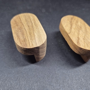 May include: Two oval-shaped wooden cabinet pulls with a natural light brown wood grain finish. Each handle features a smooth, rounded top surface and a recessed underside, designed for mounting onto furniture doors or drawers for easy gripping.