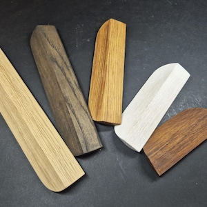 May include: Five wooden cabinet pulls in varying lengths and wood tones. The collection includes a long light oak piece, a dark walnut-stained handle, a medium-toned oak pull, a short white-washed wooden handle, and a small dark brown wooden pull.
