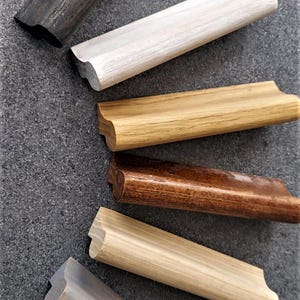 Può includere: Sei campioni di battiscopa in legno di diversi colori e finiture. I campioni sono disposti in fila dall'alto verso il basso, con il campione più scuro in alto e il campione più chiaro in basso. I campioni sono tutti larghi circa 2,5 cm e lunghi 15 cm.