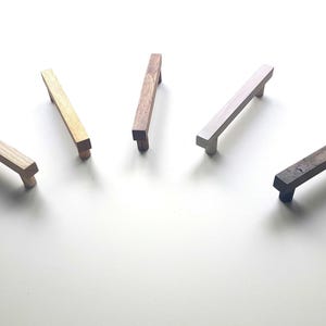 May include: Five wooden cabinet pulls in different wood tones and finishes. The pulls are rectangular with rounded edges and are approximately 3 inches long.