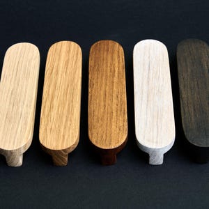 May include: Five wooden cabinet knobs in varying shades of natural wood, from light beige to dark brown and black. Each knob has a rounded top and a small, rectangular base. The knobs are arranged in a row against a dark background.