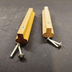 Könnte beinhalten: Zwei helle Holzgriffe für Schubladen mit rechteckigem Profil und abgerundeten Unterkanten. Jeder Griff verfügt über zwei vorgebohrte Löcher mit Metalleinsätzen. Vier silberfarbene Metallschrauben liegen bei, zwei neben jedem Griff.